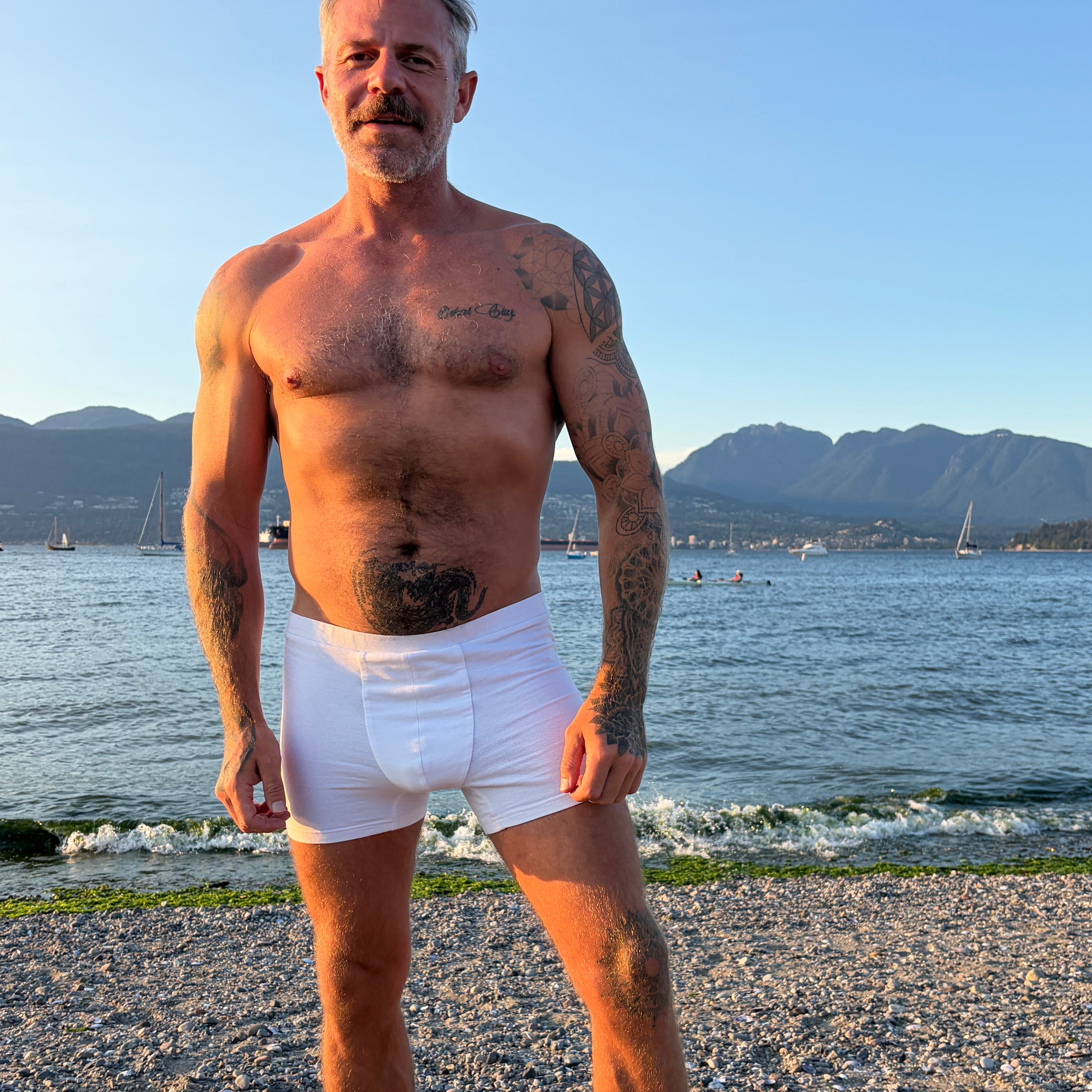 A shirtless man with gray hair and tattoos stands on a sandy beach wearing white boxer briefs, with water, distant mountains, and a clear blue sky in the background.
