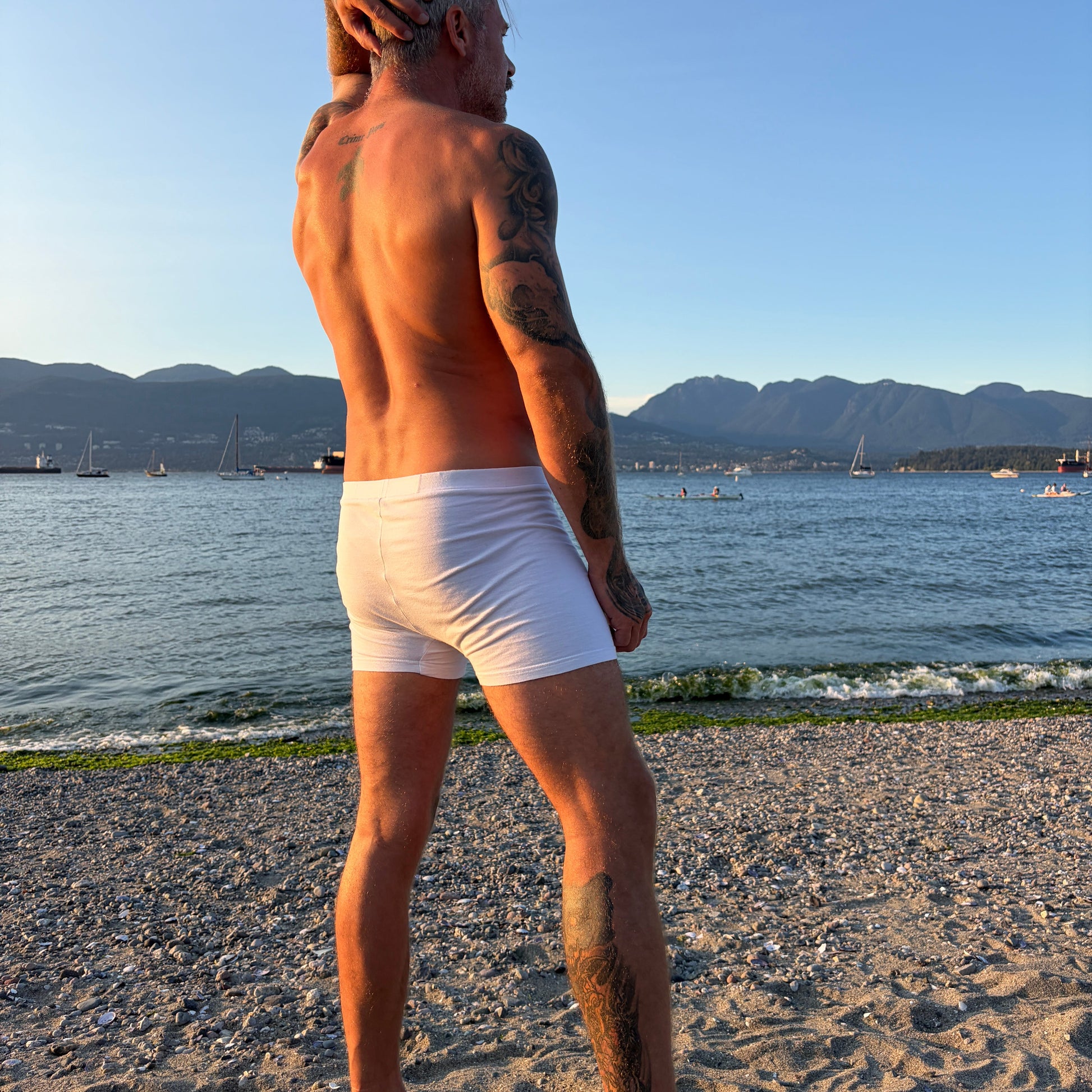 A man standing on a sandy beach facing the water, wearing white boxer briefs. The background features a calm sea, distant mountains, and a clear blue sky.