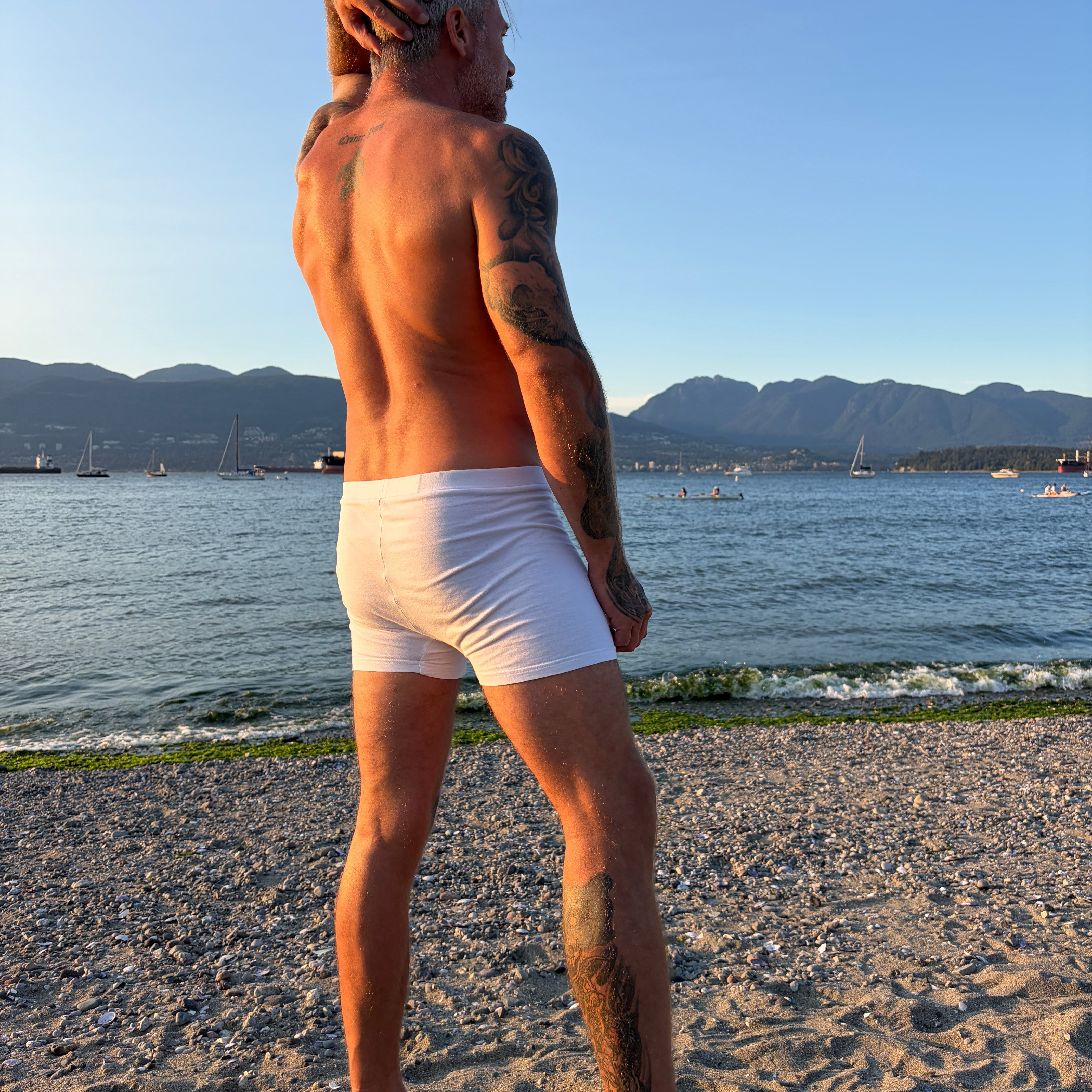 A man standing on a sandy beach facing the water, wearing white boxer briefs. The background features a calm sea, distant mountains, and a clear blue sky.