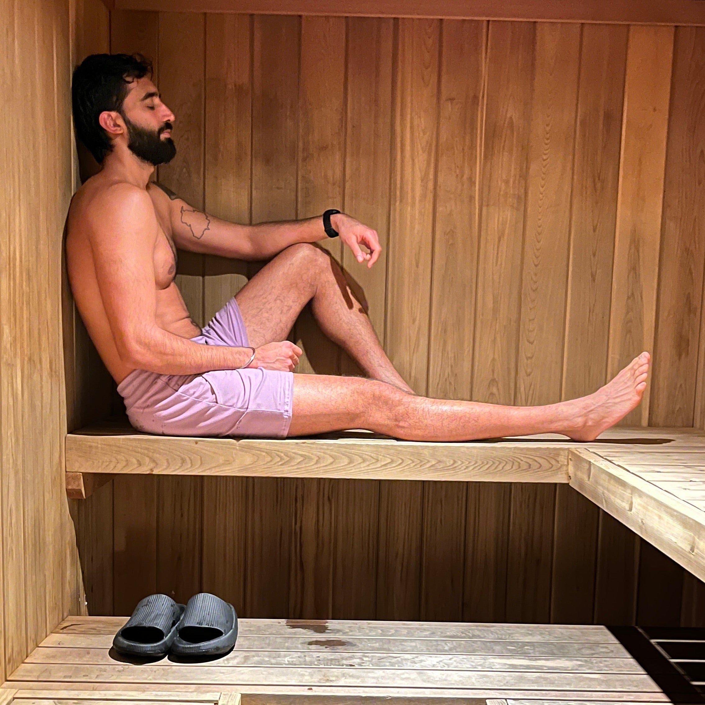 Man sitting in a wooden sauna with wooden benches, earing purple shorts and slippers on the floor.