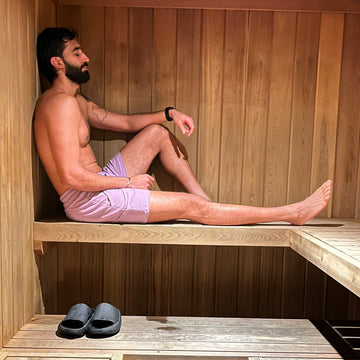 Man sitting in a wooden sauna with wooden benches, earing purple shorts and slippers on the floor.