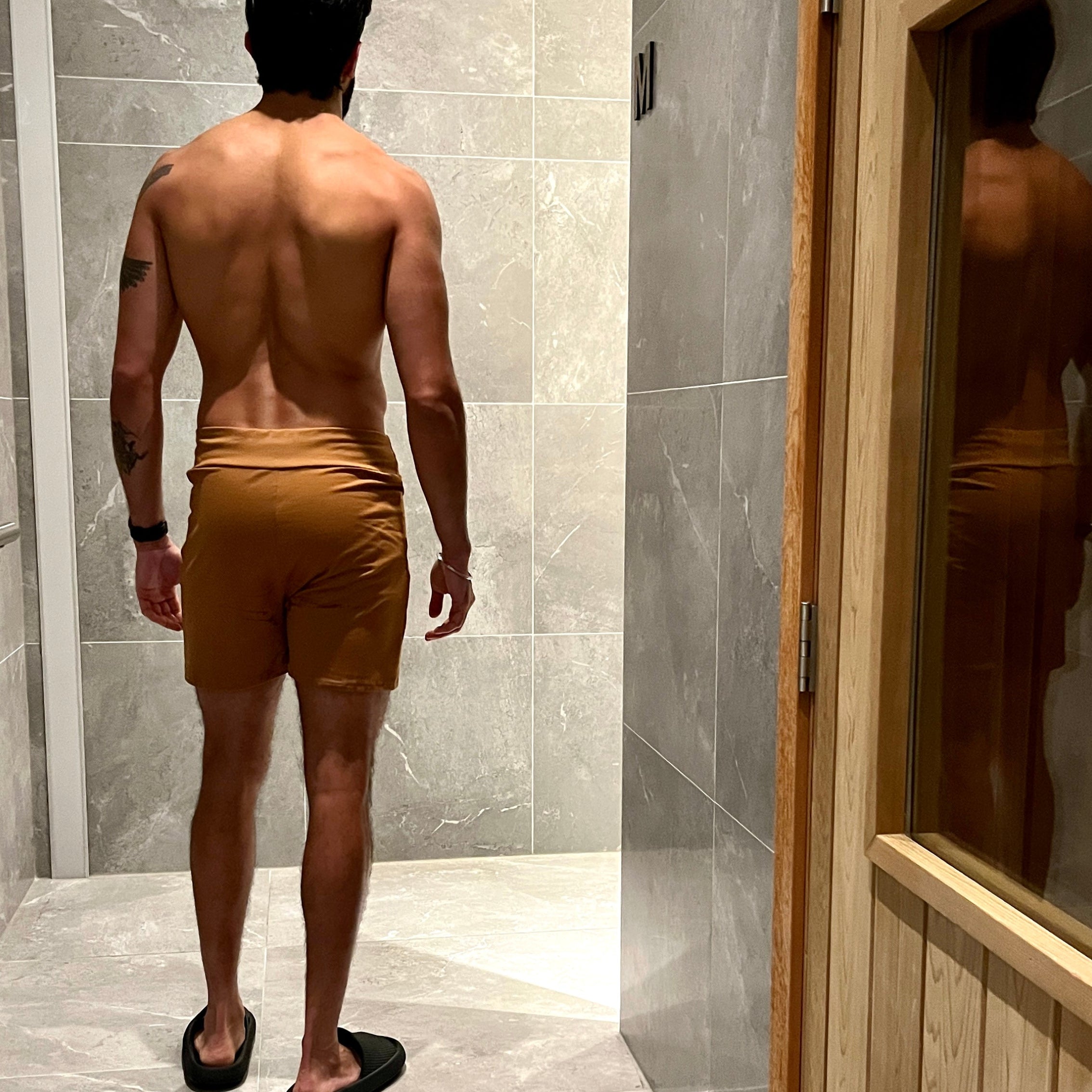 Man wearing shorts standing in a sauna change room doorway, with gray tiled walls and a wooden door.