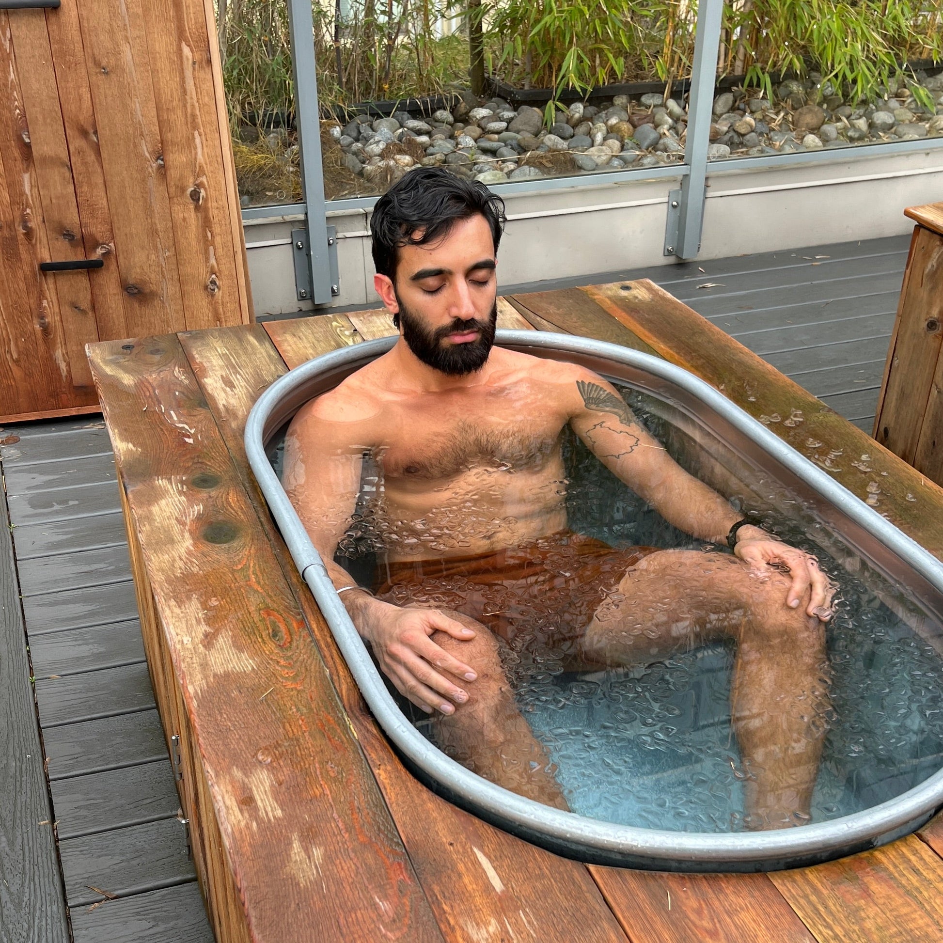 Man sitting in a metal cold plunge tub on a wooden deck with a natural outdoor setting.