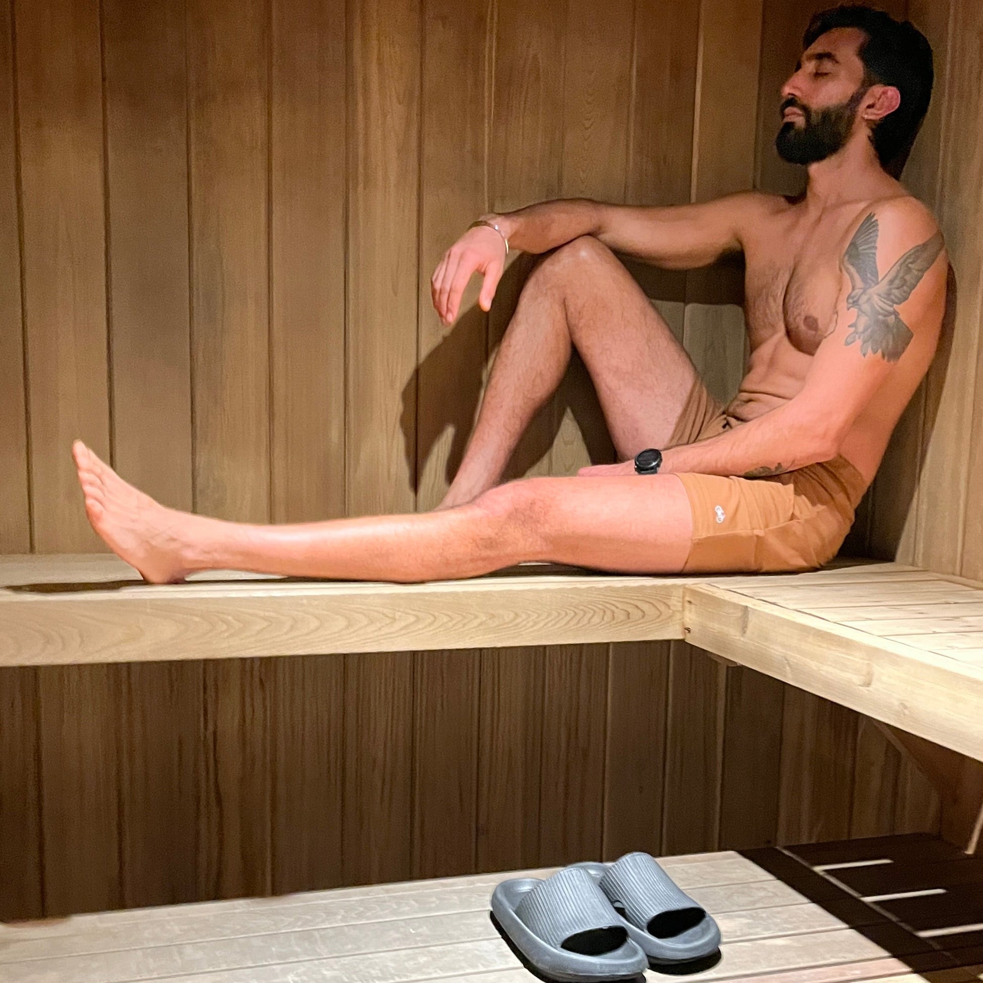 Man sitting in a wooden sauna wearing tan coloured shorts, with slippers on the floor.