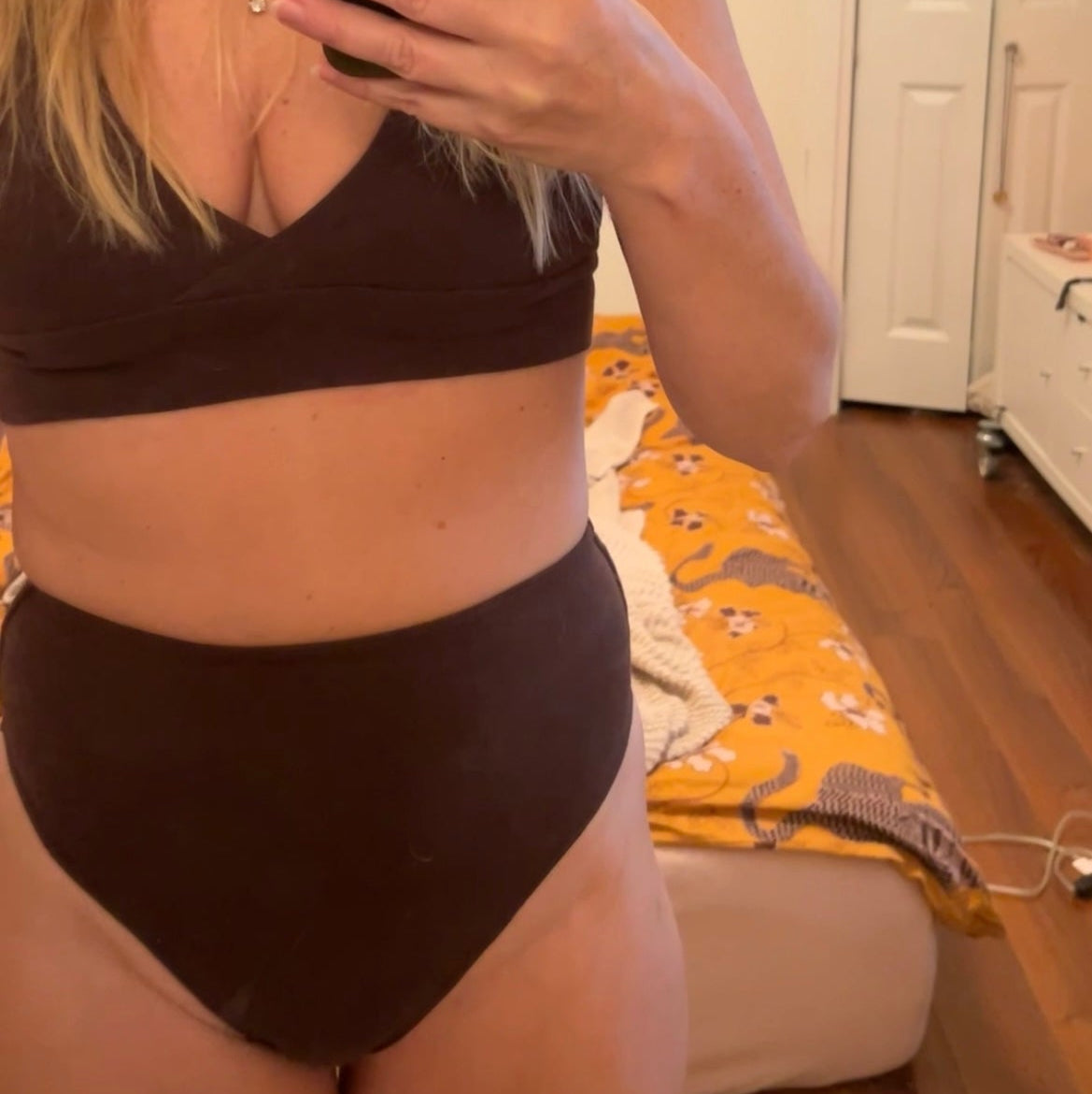 Woman taking a mirror selfie wearing a brown bikini in a room with a white door and wooden floor.