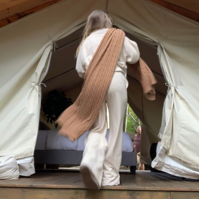 Person entering a large canvas safari tent wearing a cream outfit & a blanket over the shoulder.