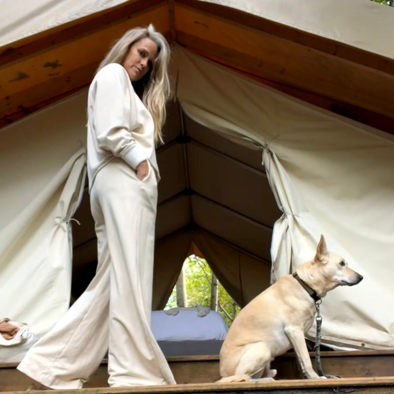 Woman in cream outfit with a dog infront of a glamping tent-like structure.