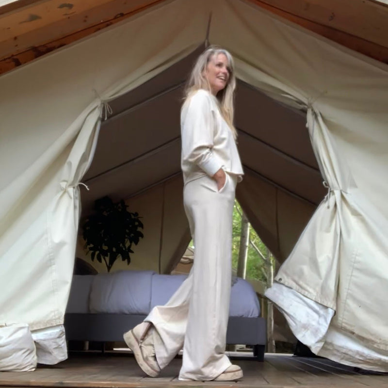 Woman standing in front of a large canvas tent with a wooden deck and trees in the background, wearing a cream outfit with her hands in her pant pockets