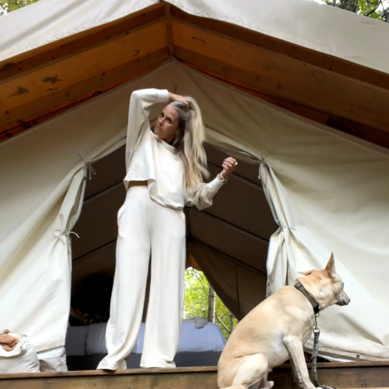 Woman in cream outfit standing in front of a glamping tent with a matching dog sitting.