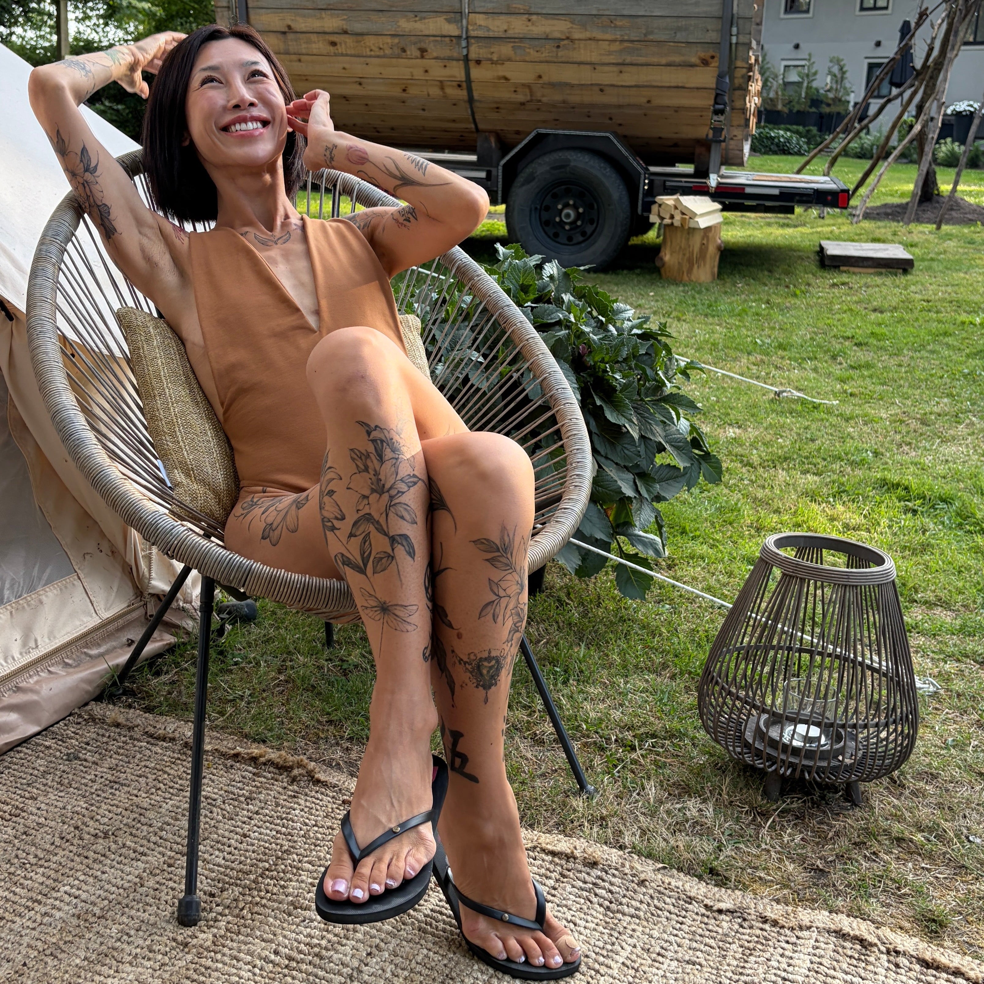 A woman with tattoos sits in a woven outdoor chair on a patio, wearing a tan bodysuit and black flip-flops, with a tiny wooden trailer and trees in the background.