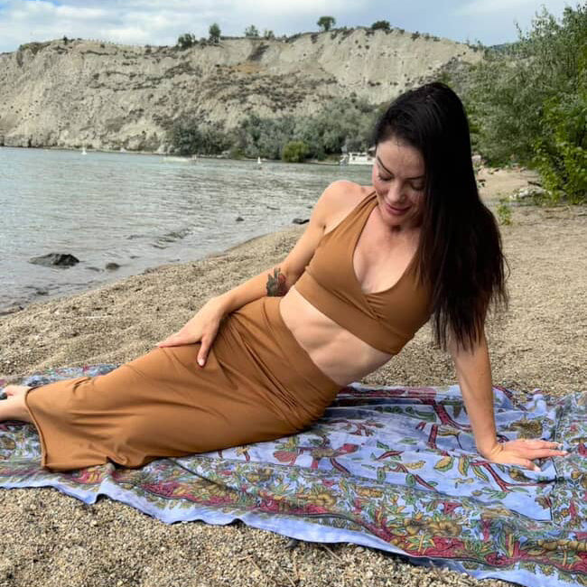 Woman in a brown outfit lying on a colorful blanket by a body of water.