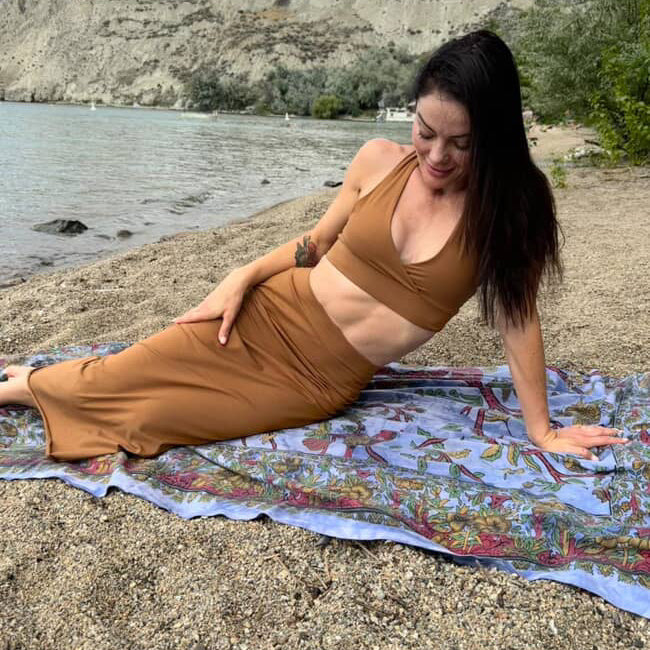 Woman in a brown outfit lying on a colorful blanket by a body of water.