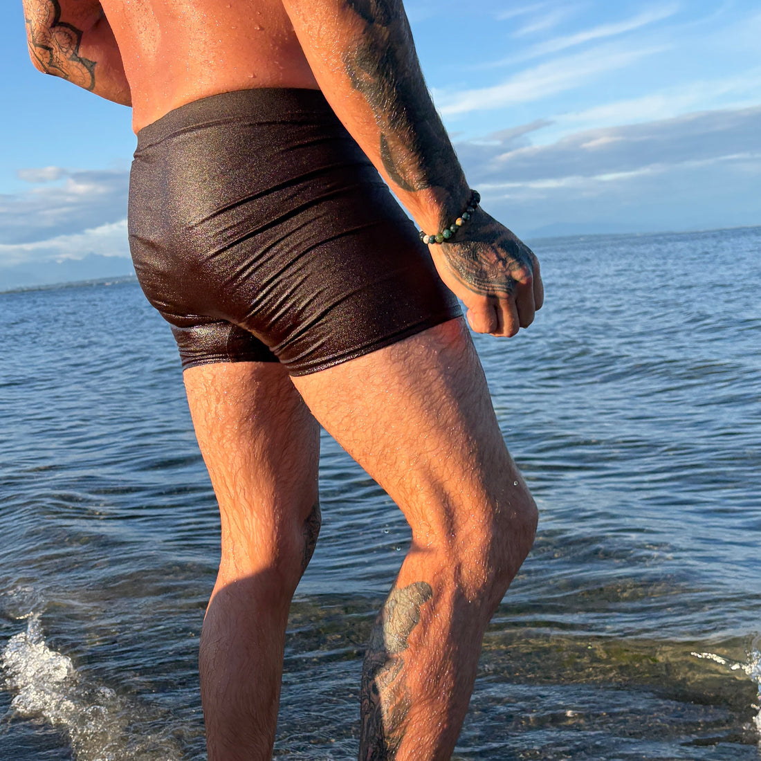 Man walking on a beach wearing dark brown swim shorts.