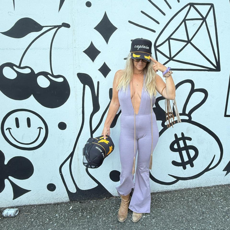 Downtown Betty in a Lavender Fields Mansion Playsuit posing in front of a graffiti-covered wall wearing a Captain Boho Boss Trucker Hat with embroidered Gold Oak Leaves