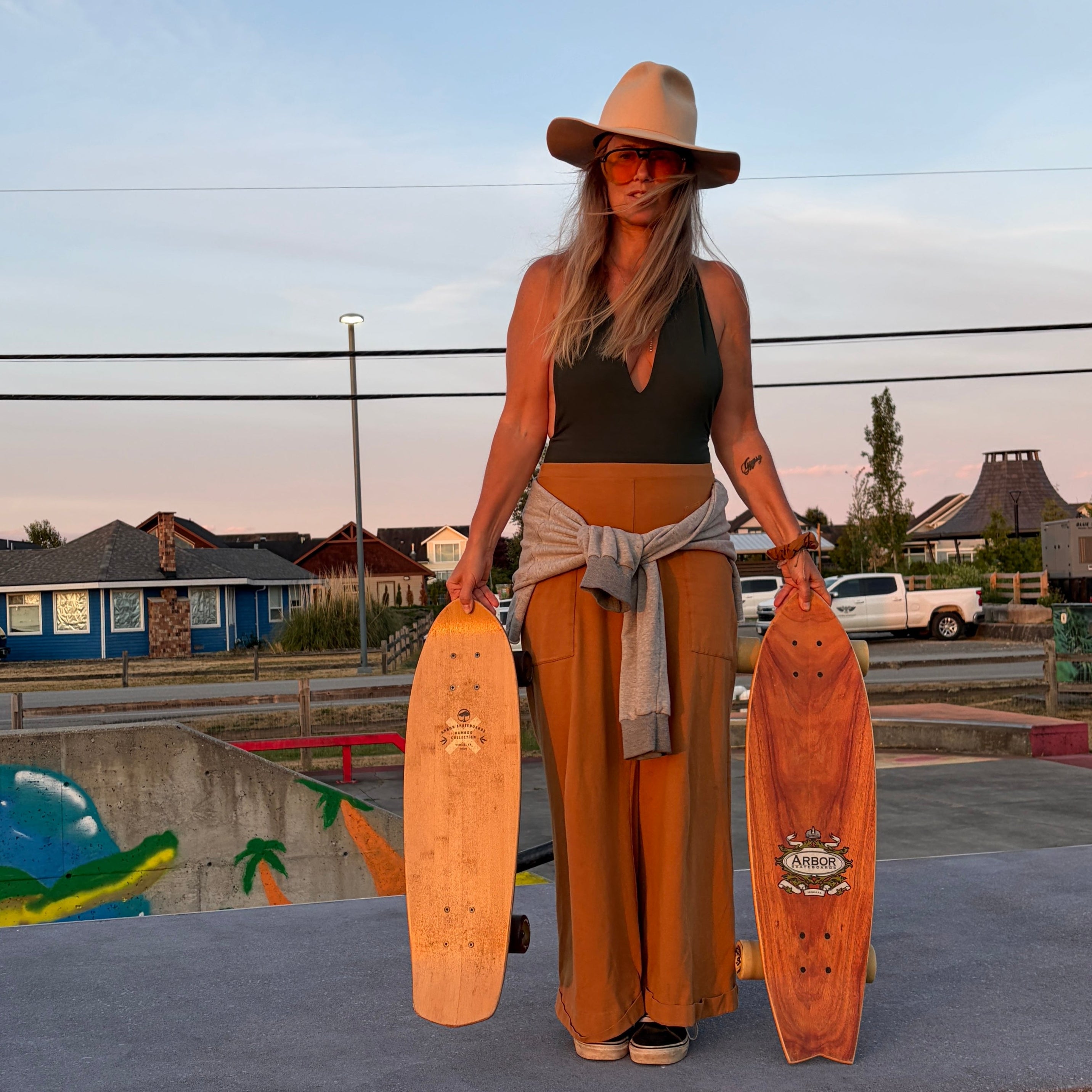 A woman wearing a wide-brimmed hat, a dark top, and wide-leg tan pants stands at an outdoor skatepark holding two longboards, one in each hand.