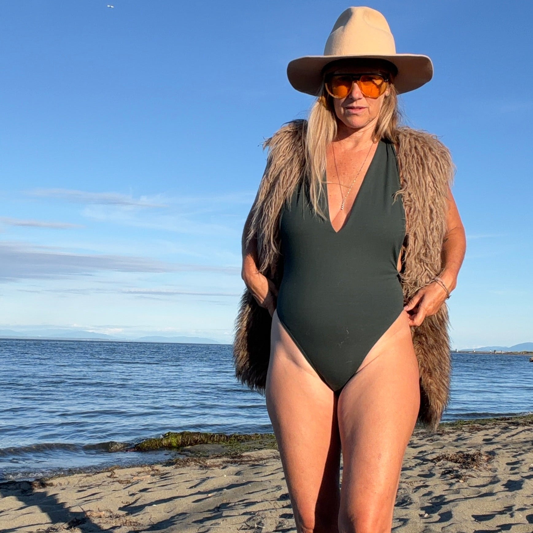 A woman stands on a sandy beach wearing a dark green one-piece swimsuit, a tan wide-brim hat, and a brown faux-fur vest, with the ocean and blue sky in the background.