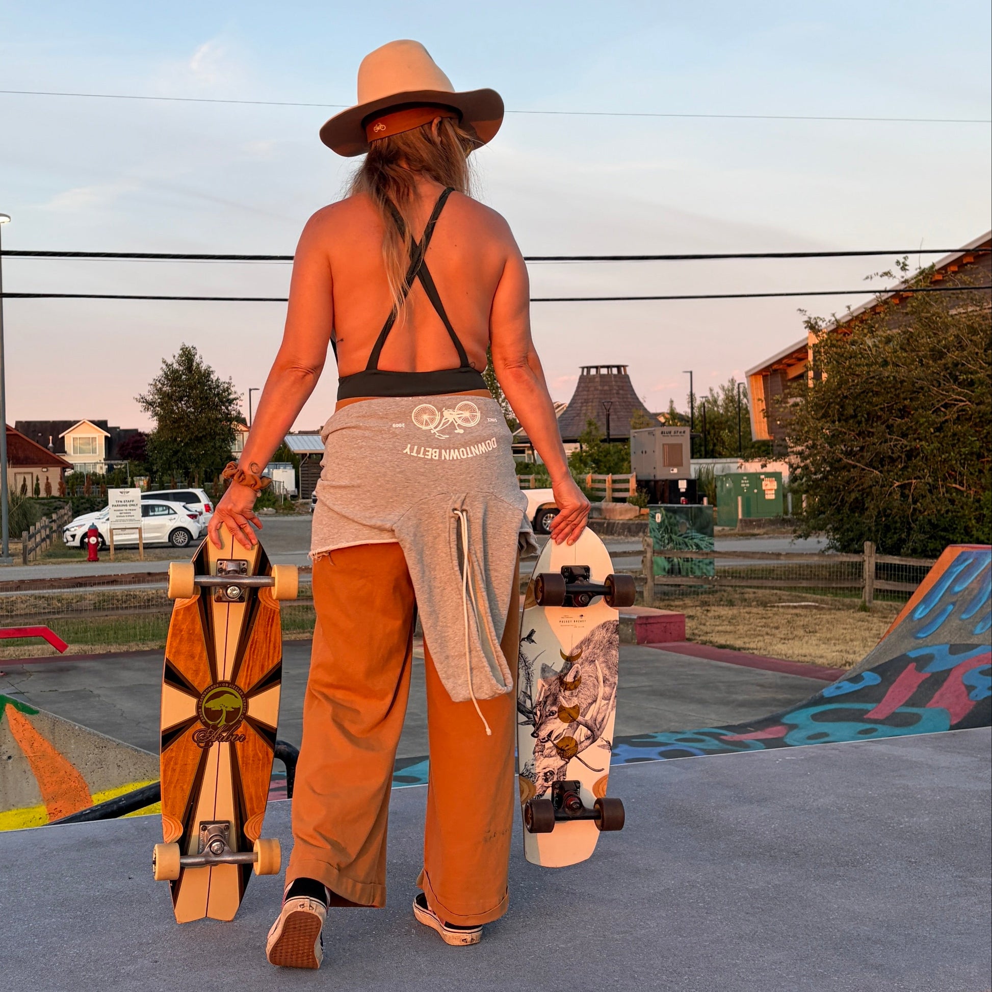 A woman stands at a skatepark holding two skateboards, viewed from behind. She is wearing a wide-brimmed straw hat, a dark green strappy top, beige wide-leg pants, and sandals.