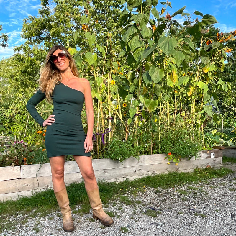 wearing a one-arm green mini dress with tan boots standing in a garden with sunflowers & greenery