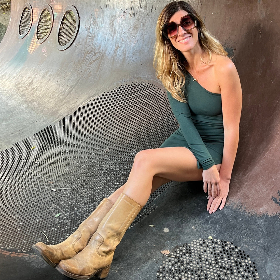 Woman in a green dress sunglasses & boots, standing inside a large rusty sculpture with a natural background