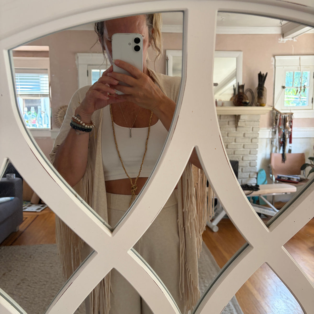 A woman takes a mirror selfie in a cozy room, wearing a beige fringe cardigan over a white crop top and matching beige pants. The photo is framed by a white decorative mirror with a geometric lattice pattern.