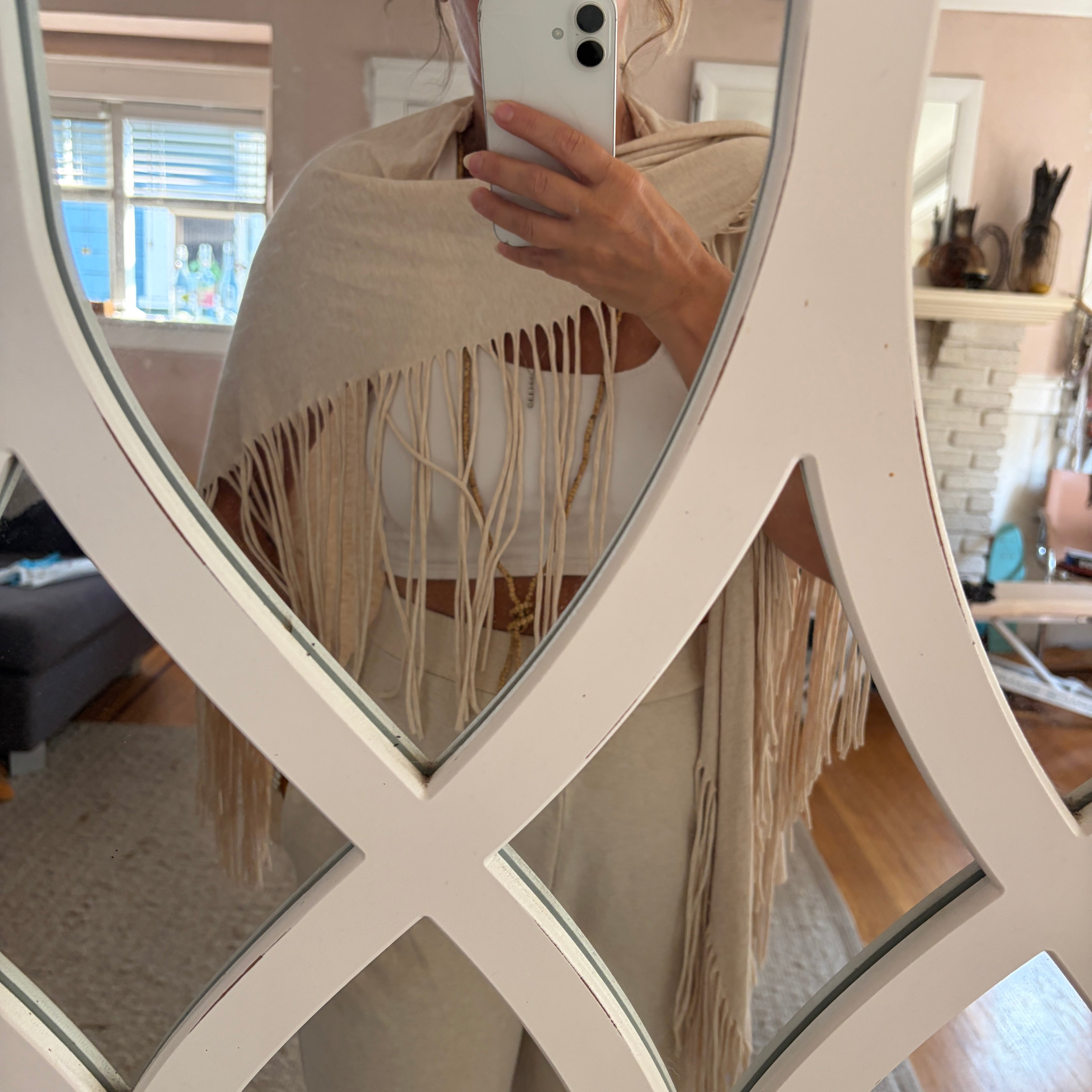 A person taking a mirror selfie in a home setting, wearing a cream-colored fringed shawl draped over their shoulders. The background shows a cozy living room with a fireplace and wooden floors, framed by a large white decorative mirror.