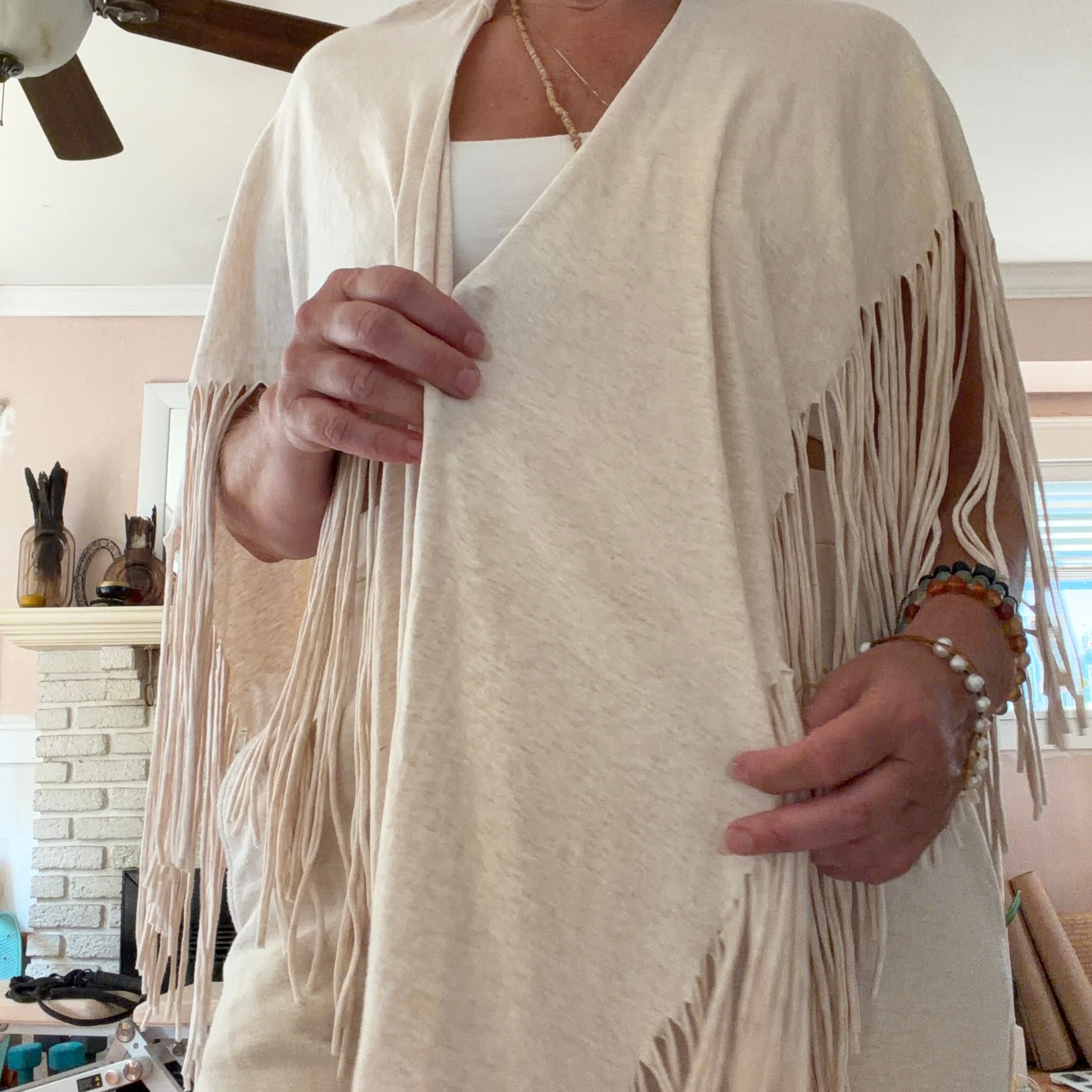A person wearing a cream-colored fringed wrap or poncho over a white top, standing in a home interior with a fireplace and ceiling fan visible in the background.