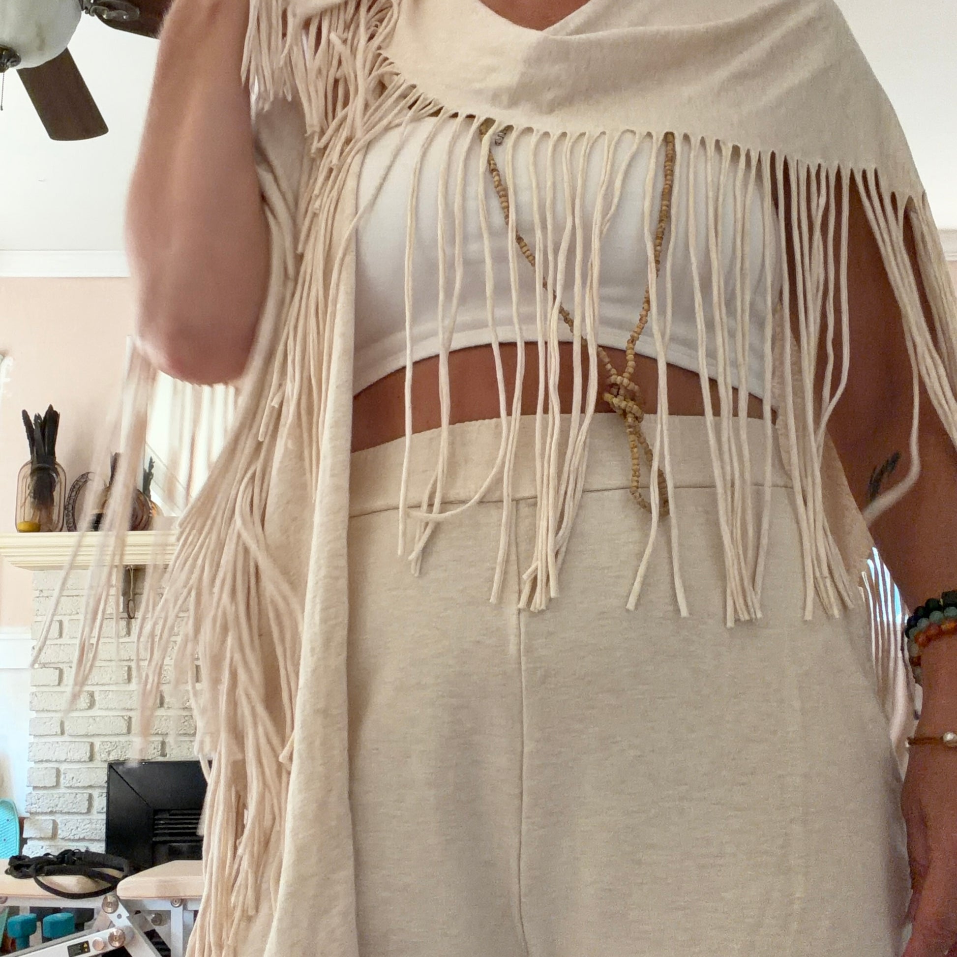 A person wearing a cream-colored fringed shawl over a white crop top and matching cream pants, standing in a home interior.