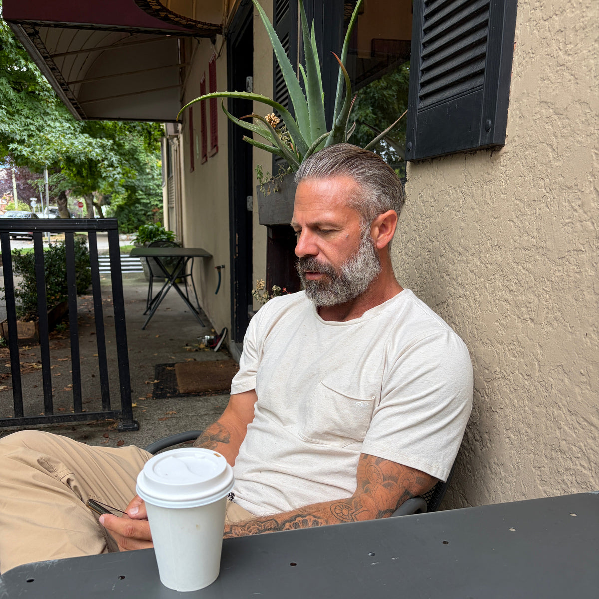 A man with a gray beard and tattoos sits at an outdoor cafe table wearing a white crewneck t-shirt and beige pants, with a white paper coffee cup in the foreground.