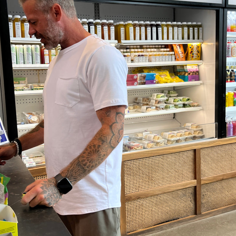 Man standing at a counter in a white T-Shirt in a health store with shelves stocked with fresh pressed juiced & sandwiches
