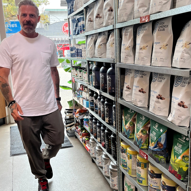 Man wearing a white Tshirt, pants & runners, standing in a supplements store aisle with shelves stocked with various protein poweders.