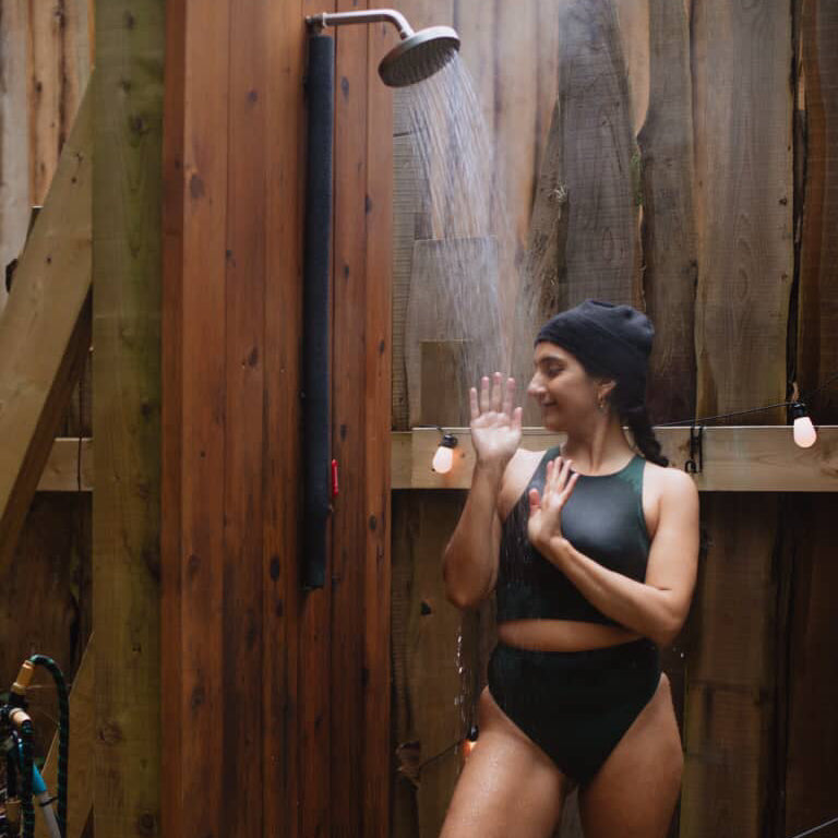 Person in a dark green natural fiber  sauna suit standing under an outdoor shower with wooden walls.