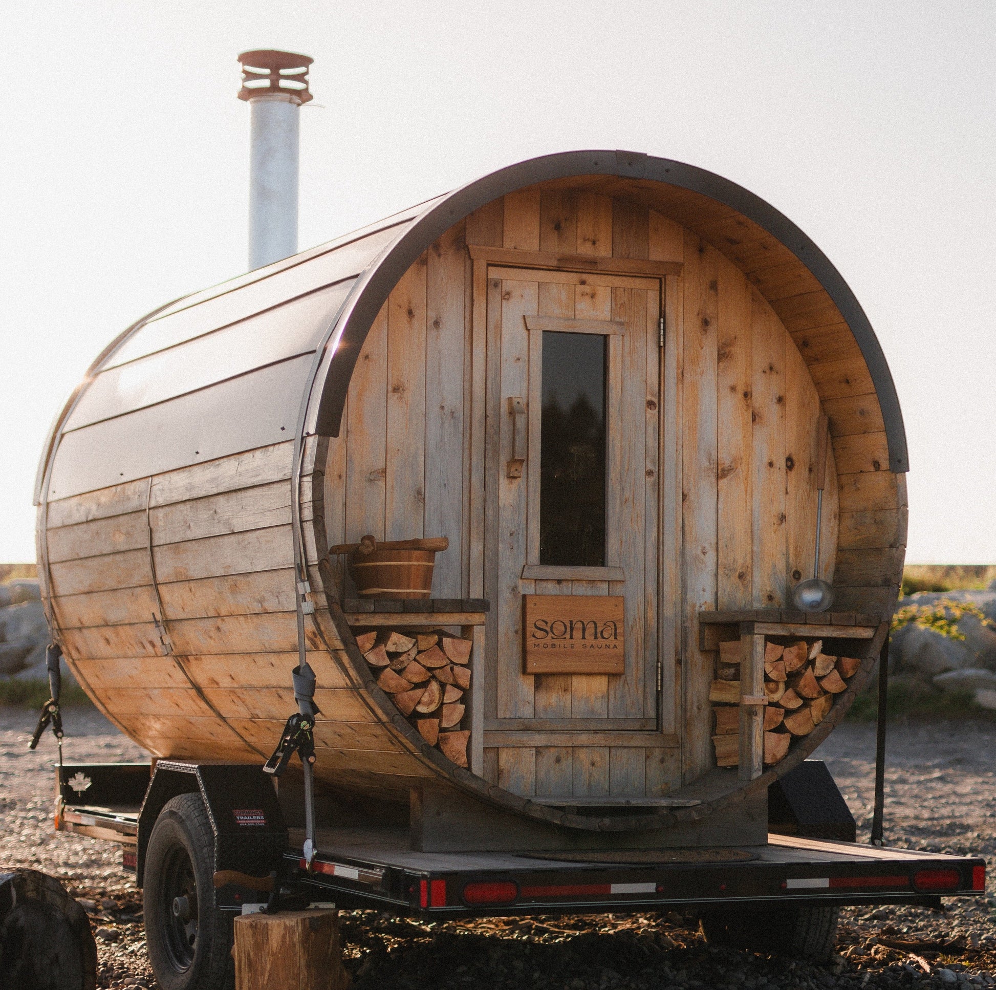 A small wooden sauna cabin on a trailer, photographed outdoors on a gravel surface with rocks and a pale sky in the background. The cabin has curved wood walls, a front door with a window, stacked firewood visible on the sides, and a metal chimney stack.