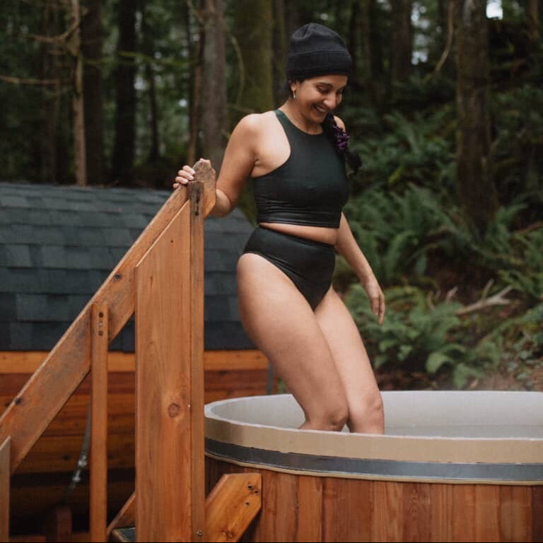 Person in a dark green natural fiber swimsuit and beanie stepping out of a hot tub in a forest setting